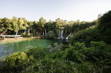 Bereketli yeşil çevresiyle Kravice şelalesi ve berrak turkuaz su güneşli bir günde rahatlama ve açık havada keşif için sakin ve pitoresk bir ortam yaratır..