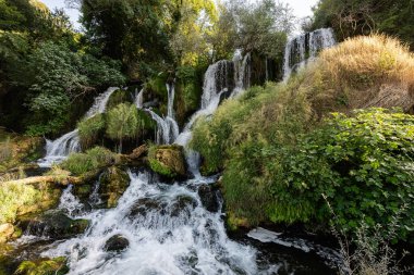 Serene Kravice şelalesi, gür orman yaprakları arasında yosunlu kayaların üzerinde çağlayarak akan su ve canlı yeşillikle sakin bir doğa sahnesini tasvir ediyor..