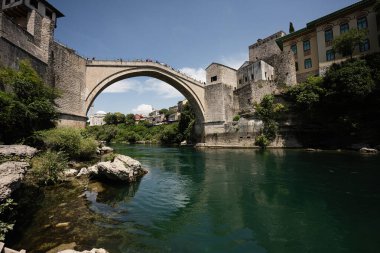 Bosna 'nın Mostar kentindeki ikonik Stari Köprüsü, güneşli bir günde tarihi mimari ve canlı yeşillikler arasında Neretva Nehri üzerinde uzanır..