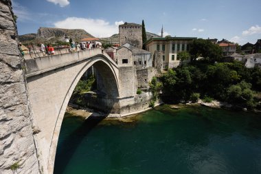 Bosna-Hersek 'in Mostar kenti - 15 Temmuz 2024: Turistler, Mostar' ın berrak nehre bakan ve tarihi mimari ve yemyeşil alanlarla çevrili ikonik Stari Most köprüsünü keşfettiler.