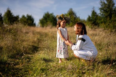 Bir anne ve kızı güneşli bir çayırda neşeli bir anı paylaşıyor. Kadın diz çöker, çocuğunun ellerini tutarken genç kız çiçekli bir taç giyer..