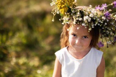 Çiçekli bir taç takan genç bir kız güneşli bir çayırda duruyor ve masum bir anı ve doğanın güzelliğini yakalıyor. Neşeli ifadesi çocukluk sevincini ve doğayla bağlantısını vurguluyor..