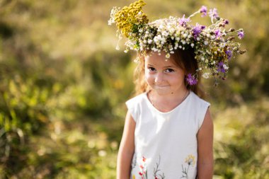 Çiçekli bir tacı olan genç bir kız, güneşli bir çayırda durup, masumiyetin, doğanın ve çocukluk sevincinin özünü yakalarken hafifçe gülümser..