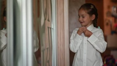 Genç bir kız aynanın önünde beyaz gömleğini ilikliyor. Okul ya da özel bir etkinlik için hazırlanıyor. Bu görüntüler aile, çocukluk veya okula dönüş videoları için mükemmel..