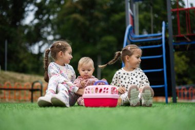 Üç genç kız kardeş dışarıda oyun parkının yanındaki çimlerin üzerinde oturarak eğleniyorlar. Takım çalışması ve dostluk gösteriyorlar, neşeli ve kaygısız bir atmosfer yaratıyorlar..