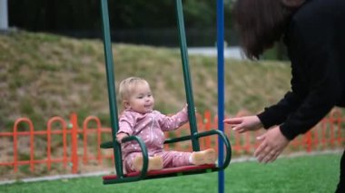 Mutlu bir kız bebek, parkta annesi tarafından salıncakta sallanmaktan zevk alır. İleri geri sallanırken gülümser ve güler..