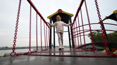 Genç bir kız güvenli bir şekilde oyun parkı köprüsünde yürüyor. Kamera hareketlerini takip ediyor, çocukluğun neşesini ve özgürlüğünü yansıtan hareketli ve çekici bir çekim yaratıyor..