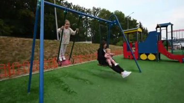 Bir anne, kızlarını salıncakta sallamaktan hoşlanır. Aile, çocukluk ve açık hava eğlencesinin iç açıcı bir sahnesi..