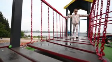 Küçük bir kız, oyun parkındaki kırmızı ve siyah metal bir köprüde dikkatle yürüyor. Denge için kırmızı zincir korkuluklara tutunur. Islak ahşap tahtalara basar.. 