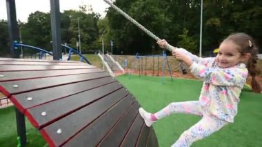 Genç bir kız oyun sahasında eğlenirken halat köprüsünde oynuyor, tasasız bir ruh ve mutluluk gösteriyor. Çocukluk temaları, oyunculuk ve açık hava eğlencesi için mükemmel..