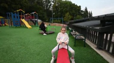 Küçük bir kız gülüyor ve oyun parkında kırmızı bir tahterevallide oynuyor. Arka planda ailesi parktaki günün tadını çıkarıyor..