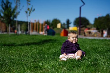 Mor ceket giyen şirin bir kız çocuğu, uzaktaki bir oyun parkında güneşli bir açık hava parkında yeşil çimlerin üzerinde mutlu bir şekilde oturuyor..