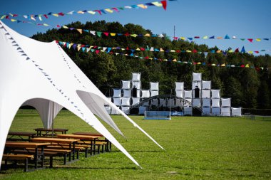 Güneşli bir günde renkli flamalı bayrakların altında yeşil bir alanda modern bir sahne ve büyük beyaz çadırların yer aldığı bir açık hava etkinliği alanı..