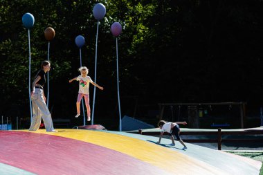 Çocuklar oyun parkında büyük, renkli bir zıpzıp yüzeyinde açık havada eğleniyorlar. Açık gökyüzünün altında canlı balonlarla dolu canlı bir sahne..