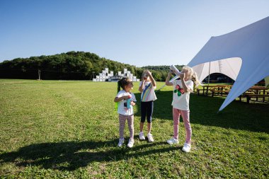 Üç çocuk, çimenli bir arazide oyuncak dürbünle keşif yapıyormuş gibi yaparak güneşli havayı seviyor. Arka planda beyaz bir çadır ve ağaçlar görülebilir..