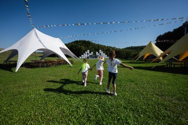 Üç neşeli çocuk bir açık hava festivalinde çimenli bir arazide koşuyor. Parlak çadırlar ve bayraklar açık mavi gökyüzünün altında şenlikli bir atmosfer ekler..