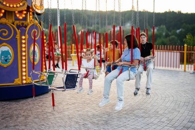 Çocuklar ve yetişkinler de dahil olmak üzere bir grup insan güneşli bir günde lunaparkta eğlenceli bir atlıkarınca gezintisi yapıyorlar..