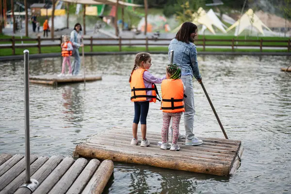 Can yeleği giyen iki çocuk, yetişkin gözetiminde bir gölette rafting yapıyorlar. Sahne bir parkta geçiyor, yaz gününde güvenli ve maceralı bir ortam sağlıyor..