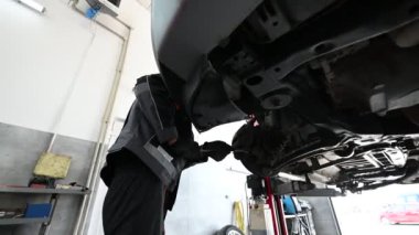 Üniformalı bir tamirci arabanın altından arka dingil üzerinde çalışırken görülüyor. İngiliz anahtarı kullanıyor ve eldiven giyiyor..