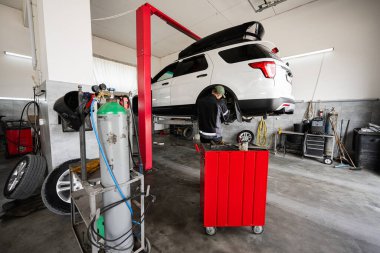 Bir tamirci profesyonel bir oto tamirhanesinde yükseltilmiş beyaz bir Amerikan cipini tamir ediyor. Araçlar ve ekipmanlar görünür olup, otomotiv atölyesinde tipik bir günü vurgular..