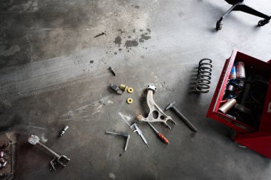 Otomotiv aletleri ve parçaları garajın beton zeminine, açık bir alet çantasının yanına saçılmış. Sahne, el işçiliği ve mekanik tamirat hissi uyandırıyor..