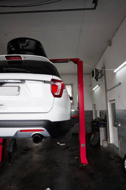 Modern bir oto tamirhanesinde beyaz bir Amerikan arabası hidrolik asansörle kaldırıldı. Araç bakım ve onarım için konumlandırıldı. Otomotiv hizmetlerinde kullanılan yüksek teknolojili ekipmanları gösteriyor..