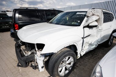 Ön ve yan tarafında büyük hasar olan beyaz bir SUV muhtemelen çarpışmadan sonra bir otoparkta duruyor. Hava yastığı açılmış, kazanın şiddetini gösteriyor..