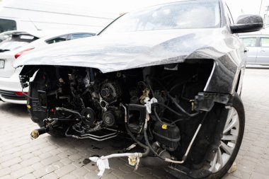 Ön tamponu hasar görmüş ve motor parçaları ortaya çıkmış bir arabanın yakın çekimi. Araç açık hava otoparkında, bir kazanın sonrasını vurguluyor..