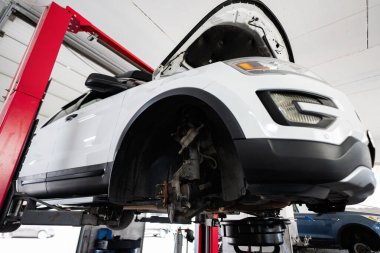 Beyaz bir Amerikan SUV bir garajın içinde hidrolik bir asansörle bakım ve onarım için hazır hale getirilir. Görüntü otomotiv onarım hizmetlerini ve aracın mekanik yönlerini vurgular.