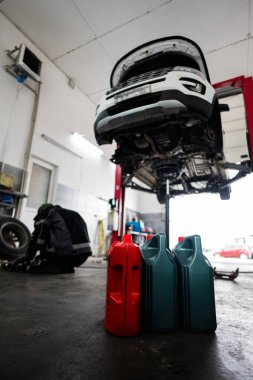 Bir otomobil tamirhanesinde hidrolik asansörde bir araç yükselir. Petrol konteynırları ön planda, garajdaki rutin araba bakım ve servis işlerini vurguluyorlar..