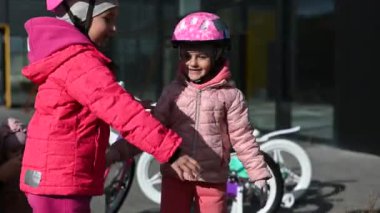İki mutlu küçük kız sarılıp baş parmağını kaldırıyor. Pembe, gül rengi ceketler ve bisiklet kaskları giyiyorlar. Arka planda bir denge bisikleti görünür.