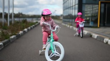 İki mutlu küçük kız modern bir binanın yakınındaki asfalt bir yolda bisikletlerine biniyor. Pembe, deniz mavisi ceketler ve miğferler giyiyorlar ve kameraya bakıyorlar.. 