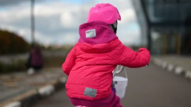 İki küçük kız modern bir binanın yanındaki kaldırımlı bir yolda bisiklet sürüyor. Bir kız pembe ceket ve kask giyerken, diğeri açık yeşil ceket ve kask giyiyor..