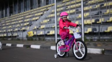 Küçük mutlu bir kız stadyumun yanında bisikletiyle geziyor. Rüzgâr saçlarını savururken gezmekten zevk alıyor..