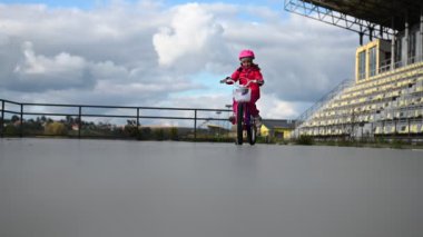 Küçük bir kız nehir boyunca asfalt bir yolda bisikletini sürüyor. Pembe bir ceket ve kask giyiyor. Sonbahar ağaçları ve bulutlu gökyüzü resim sahnesini tamamlıyor.