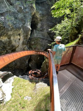 A young individual stands on a metal walkway, overlooking a spiral pathway in a lush canyon in Liechtensteinklamm, Austria. The scene exudes adventure and tranquility within a picturesque natural setting. clipart