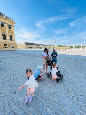 Bir aile, Avusturya 'nın Schonbrunn Sarayı' ndaki büyük sarayın bahçesinde güneşli bir günün tadını çıkarırken ebeveynler geniş kaldırım taşları bölgesindeki mimariye hayran kalırken çocuklar bebek arabasının etrafında oynuyorlar..