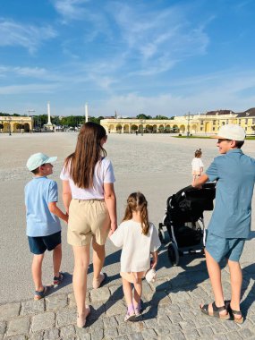 Aralarında çocuklar ve bebek arabasının da bulunduğu beş kişilik bir aile, Avusturya 'nın Schonbrunn sarayındaki geniş tarihi bir meydanda güneşli bir günün tadını çıkarıyor. Sahne, güzel bir açık hava ortamında beraberlik ve macera hissini yakalar..