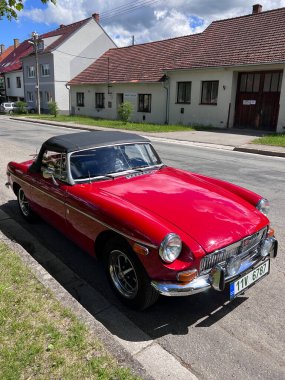 Lysice, Çek Cumhuriyeti - 28 Mayıs 2022: Güneşli bir gökyüzünün altında sessiz bir banliyö sokağına park edilmiş klasik kırmızı üstü açık araba MG B Roadster (1965). Büyüleyici klasik araç ilginç evlerin ve yemyeşil otlakların arka planında göze çarpıyor.. 