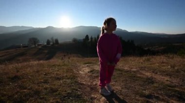 Pembe eşofmanlı genç bir kız gün batımında bir tepenin üzerinde duruyor, kollarını neşe ve özgürlük içinde kaldırıyor, dağlarla ve arka planda açık bir gökyüzüyle..