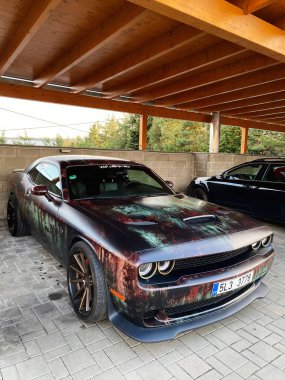 Brno, Czech Republic - April 15, 2022:  Classic muscle car Dodge Challenger Hellcat 6.2 with a faded, weathered exterior is parked under a wooden carport.  clipart