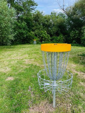 Mavi gökyüzünün altında canlı ağaçlarla çevrili yemyeşil bir parkta göze çarpan bir disk golf sepeti duruyor. Açık hava eğlencesi ve spor meraklıları için mükemmel..
