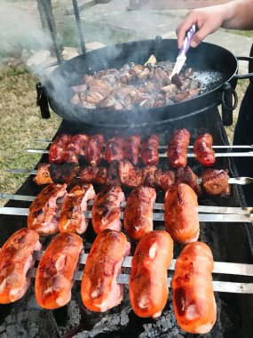 Açık hava ızgarasında cızırdayan çeşitli et ve sosislerin sergilendiği yakın plan bir çekim. Bir el, arka bahçedeki barbekü partisinin özünü yakalıyor..