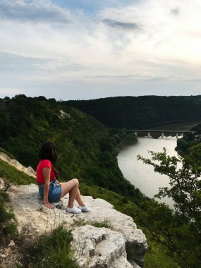 Sıradan giyinmiş bir kadın uçurumun kenarında oturur ve güzel bir nehir vadisine bakar. Manzara yeşilliklerle doludur ve gökyüzü bulutludur, sakin bir atmosfer yaratır..