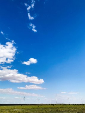 Rüzgâr türbinlerinin engin mavi gökyüzünün altında ve dağınık bulutların altında durduğu manzaralı bir manzara. Manzara, doğayla uyum içinde yenilenebilir enerji kaynaklarını yakalar..