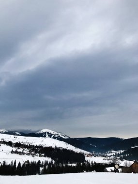 Karla kaplı tepeleri ve yoğun ormanlarla çevrili bulutlu gökyüzü ile sakin bir kış dağı manzarası. Samimi bir kulübe bu huzurlu manzaraya cazibe katıyor..