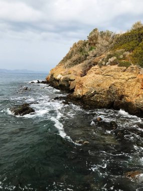 Güzel bir kayalık kıyı şeridi bulutlu bir gökyüzünün altında kıyıya vuran dalgalar, sakin doğal bir manzarayı ve huzurlu bir deniz manzarasını resmediyor..