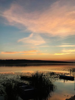 Sakin bir göl üzerinde sakin bir günbatımı manzarası ve kıyı şeridi boyunca yavaşça demirlenmiş tekneler. Gökyüzü yumuşak pastel renklerle boyanır. Huzurlu ve sakin bir atmosfer yaratır..