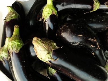 Yakın plan taze, parlak patlıcanlar üst üste dizilmiş, koyu mor renkleri ve parlak yeşil sapları sergileniyor. Organik ürünlerin ve mutfak malzemelerinin mükemmel tasviri.