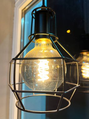 Metal kafes armatüründe parlayan bir ampul, modern endüstriyel bir tasarımı sergiliyor. Sıcak, ortam aydınlatması rahat bir atmosfer yaratıyor. Çağdaş iç mekanlar için mükemmel..